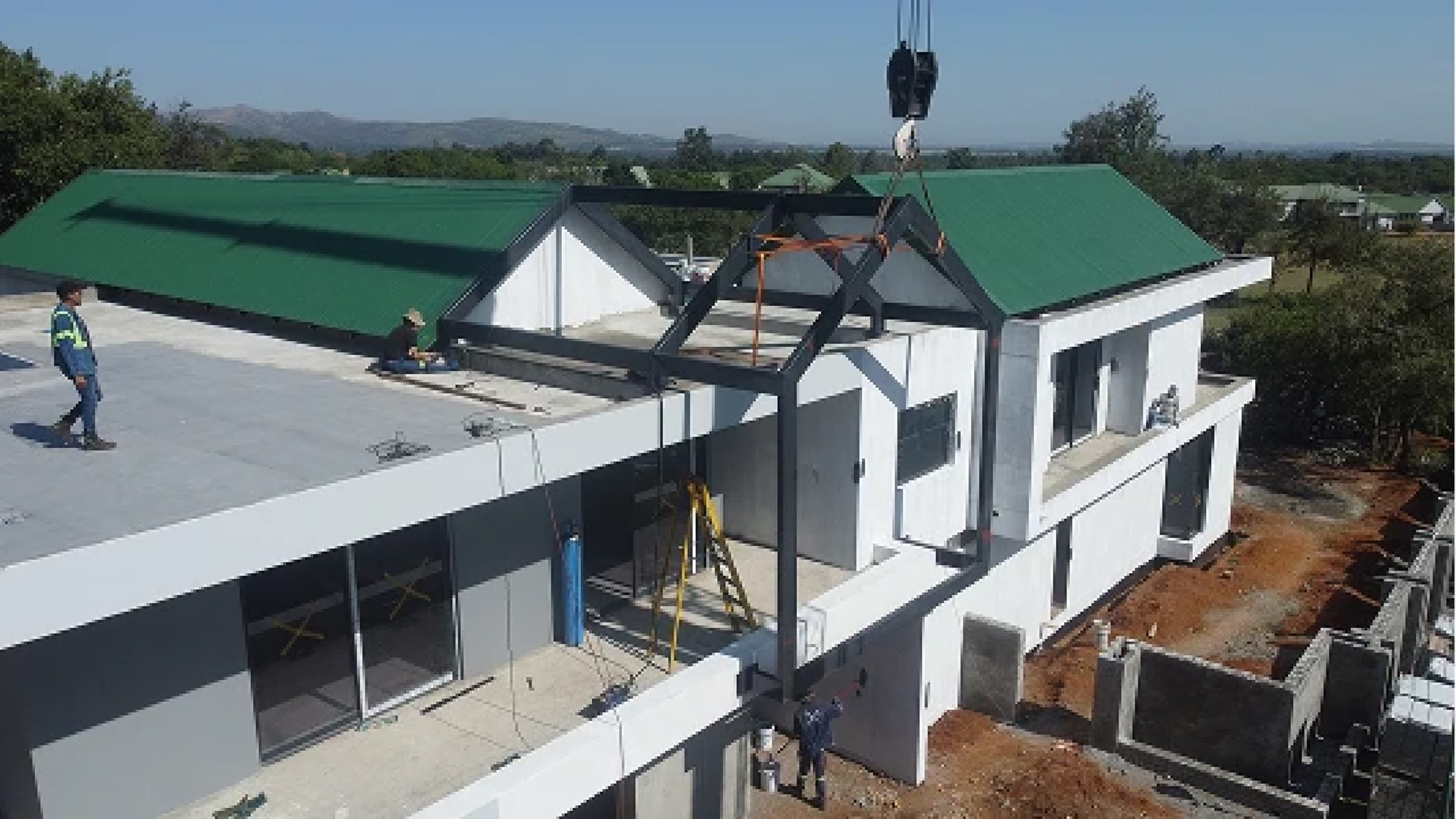 A large two-story house under construction, featuring modern architecture with green gabled roofs. A crane lifts materials, with several workers visible.