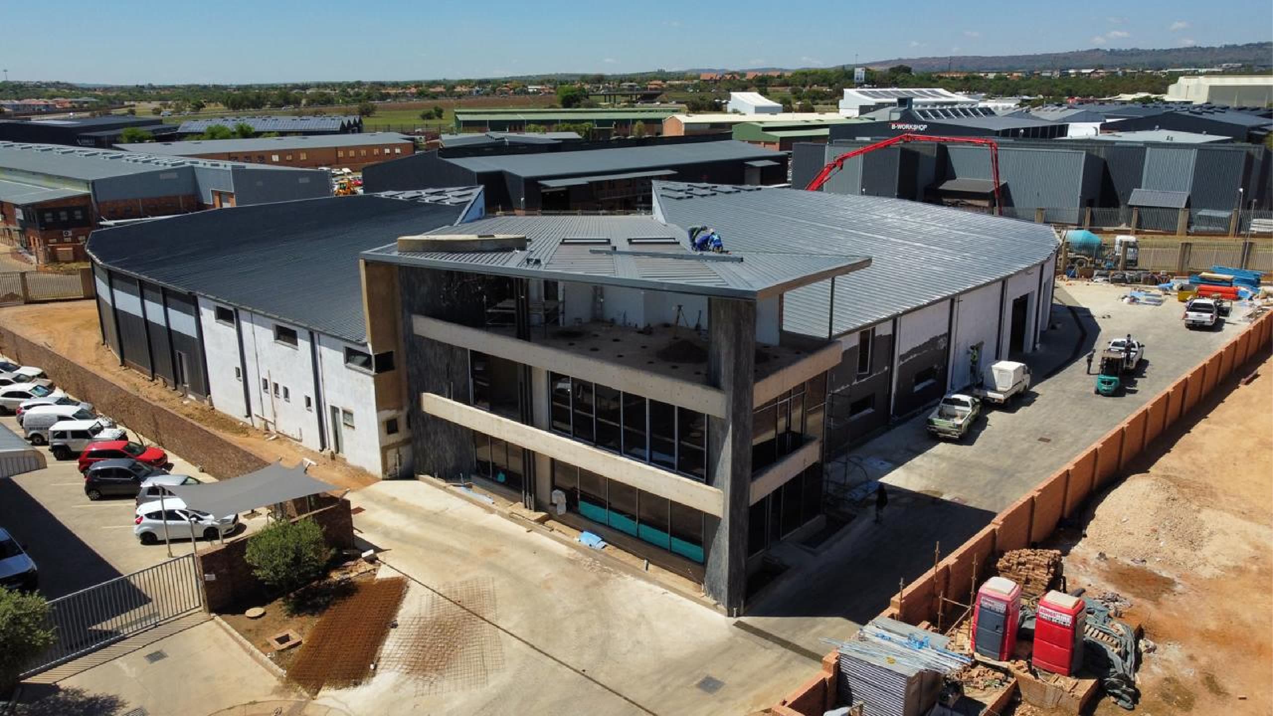 Aerial view of a modern commercial, chemical storage, and office building under construction. The building has a curved roof and large glass windows, surrounded by construction vehicles and materials.