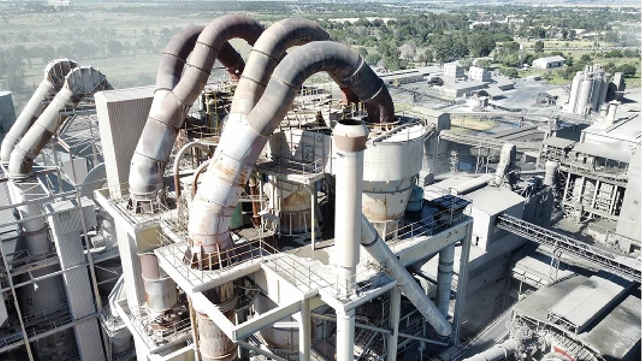 Industrial plant with large, rusted pipes and storage tanks, surrounded by machinery and greenery in the background. Aerial view shows scale.