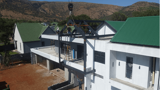 Aerial view of a white building under construction with workers installing a metal roof frame. Green-roofed houses and mountains are visible in the background.