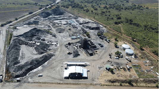 Aerial view of an industrial site with large piles of materials, machinery, and storage tanks. Surrounding landscape is green and open, under a clear sky.
