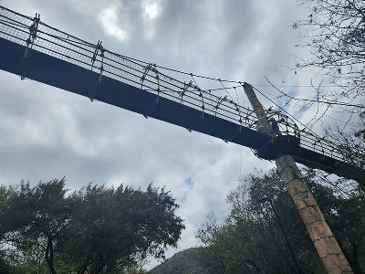 A suspension bridge stretches across the image under a cloudy sky, supported by a tall stone pillar. Trees frame the scene, conveying a sense of adventure.