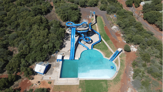 Aerial view of a water park featuring several tall, intertwining blue water slides leading into a large, clear pool, surrounded by lush greenery and paths.