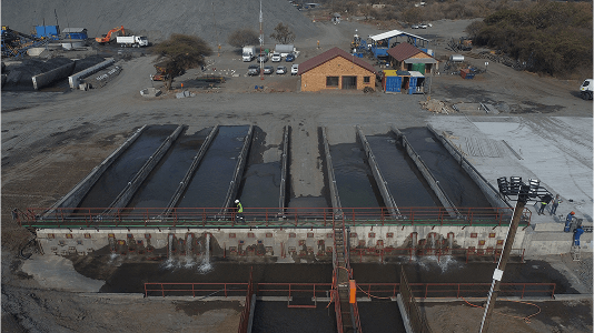 Aerial view of an industrial water treatment facility with six long channels, adjacent buildings, vehicles, and a worker in safety gear, set in a rugged area.