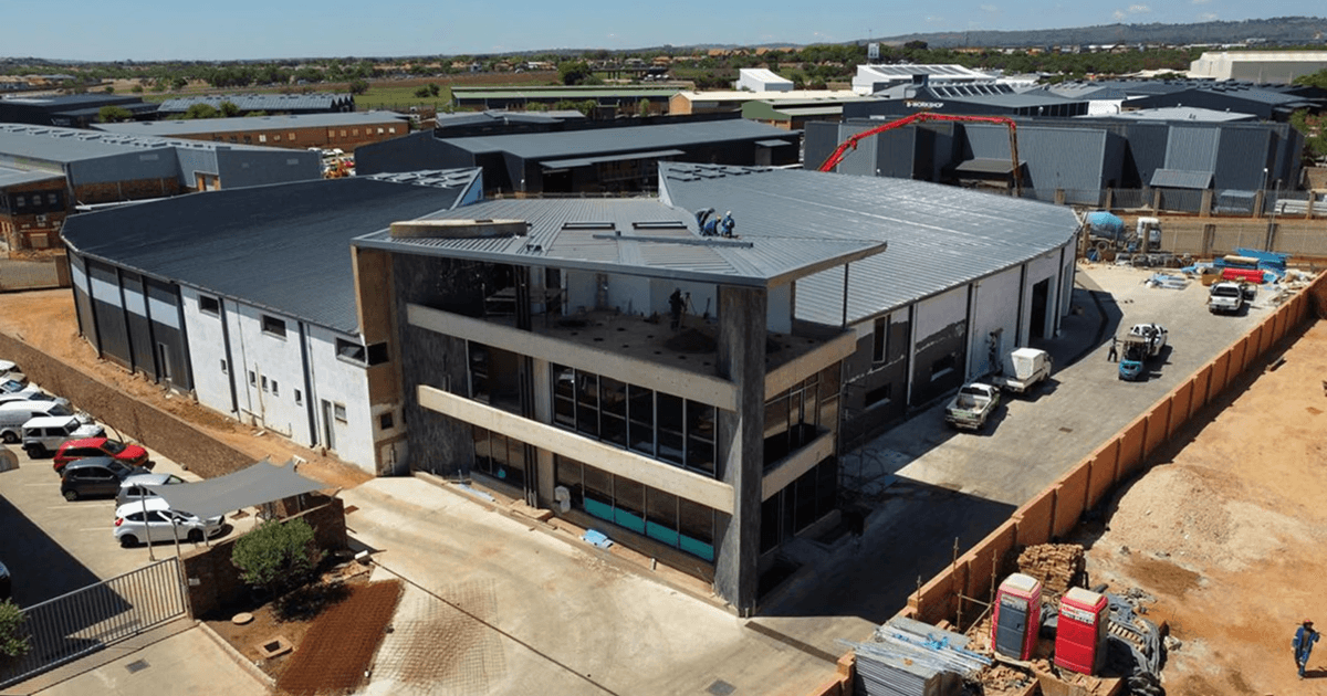 Aerial view of a modern commercial, chemical storage, and office building under construction. The building has a curved roof and large glass windows, surrounded by construction vehicles and materials.